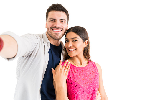 Portrait of beautiful woman showing engagement ring while boyfriend taking selfie against plain background