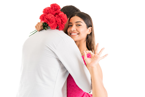 Smiling beautiful woman with flowers gesturing ok sign while embracing boyfriend over isolated background