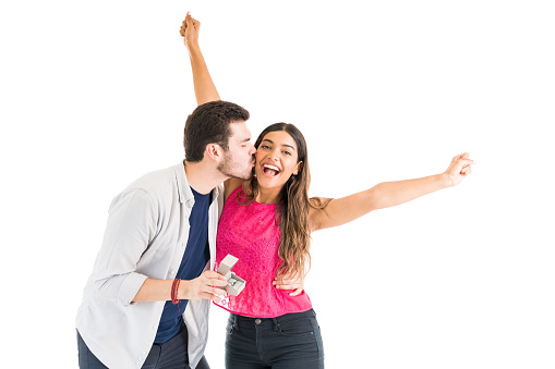 Young man holding engagement ring while kissing girlfriend standing with arms raised in excitement against plain background