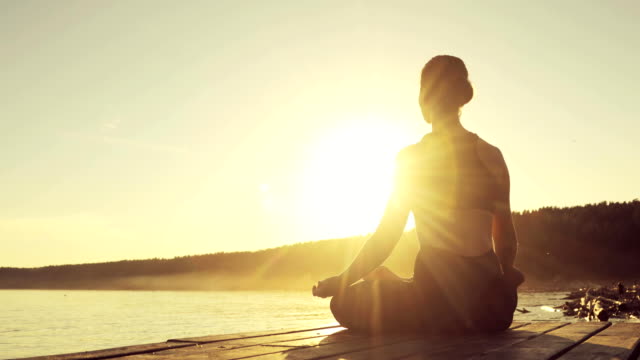 Woman is meditating in Lotus Pose sitting on pier near the river at sunset