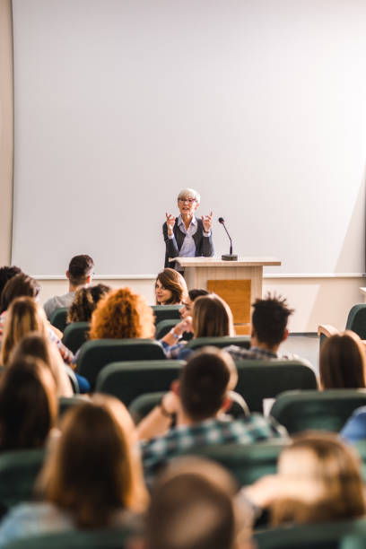 Senior teacher talking to large group of college students in amphitheater. Mature professor giving a lecture in front of projection screen at lecture hall. Copy space. teacher projector stock pictures, royalty-free photos & images