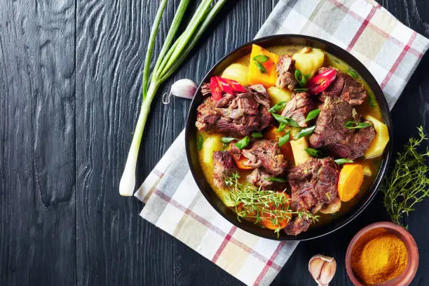 traditional Jamaican Curry Goat "u2013 slow cooked Jamaican Spiced meat and vegetables spicy Curry in a black bowl on a rustic wooden table, close-up