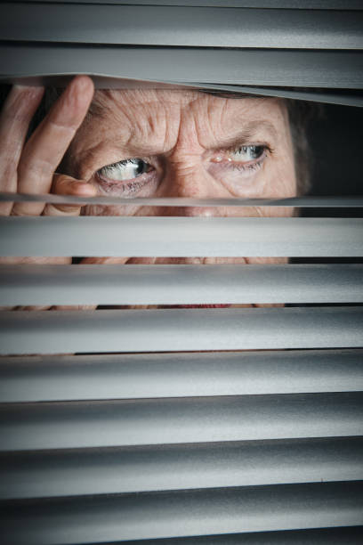 istock Wrinkled old woman peeps sideways through venetian blinds, looking scared 1161115693