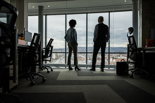 Back view of two business people standing in the office and looking through window. Copy space.