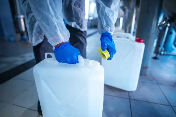 Unrecognizable person technologist in white protective suit handling acid or detergent in chemical industry. Industrial worker opening plastic canister to use chemicals.