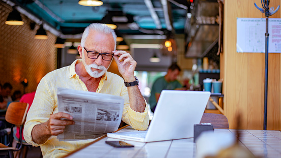 Photo of a senior man reading a newspaper wearing eyeglasses while relaxing in a restaurant during the day. Morning routine in the favorite cafe bar. Senior man in yellow shirt reading newspaper with a white laptop on the table.