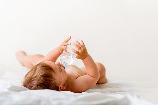 Baby toddler sits on the white bed, smiles and drinks water from plastic bottle
