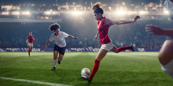 A close up image of a professional women soccer player poised with right leg back about to kick football during a soccer game in a generic floodlit stadium. The player is near opposition players and is wearing a generic red and white soccer kit. With selective focus and bokeh effects.