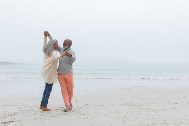 Couple dancing at the beach Front view of diverse happy senior couple smiling and dancing on the beach on cloudy day. Authentic Senior Retired Life Concept middle aged couple dancing stock pictures, royalty-free photos & images