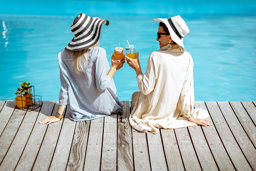 Two women in hats and shirts sitting with drinks on the poolside, back view