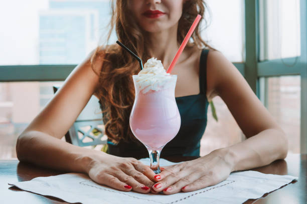 giovane bella ragazza che beve un frullato al gusto di fragola in un ristorante sullo sfondo di una finestra alla luce del giorno. la ragazza tiene un bicchiere di cocktail analcolico. - luxury milk dinner glass foto e immagini stock