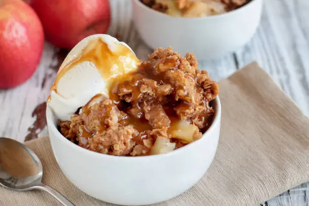 Fresh hot homemade apple crisp or crumble with crunchy streusel topping topped with vanilla bean ice cream and Caramel Sauce over rustic white table. Selective focus with blurred background.