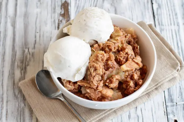 Fresh hot homemade apple crisp or crumble with crunchy streusel topping topped with vanilla bean ice cream over rustic white table. Selective focus with blurred background.