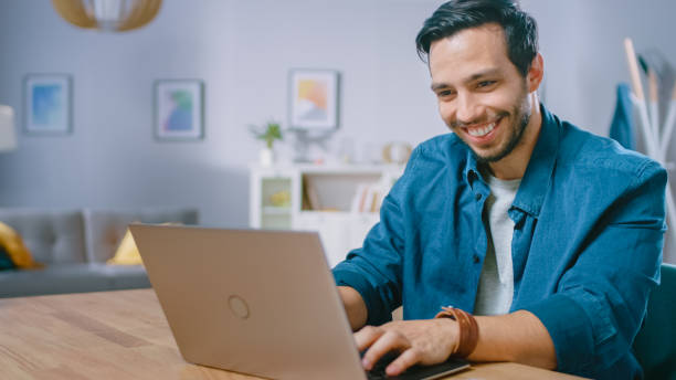 istock Portrait of Handsome Young Man Using Laptop Computer at Home, Watching and Smiling at a Content. Cozy Modern Flat done in Scandinavian Style. 1161176785