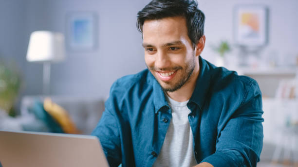 istock Portrait of Handsome Young Man Using Laptop Computer at Home, Watching and Laughing at Content. Happy Hispanic Man Works on Computer in a Cozy Living Room. 1161176806