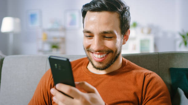 istock Happy Young Man Uses Smartphone while Sitting on a Sofa at Home. Man Browses Through Internet, Watches Videos and Uses Social Networks at Home. 1161176867