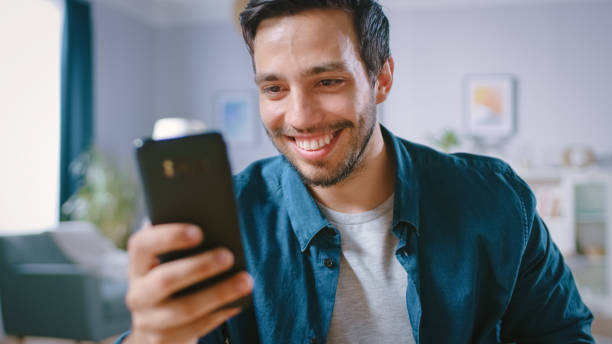 istock Handsome Smiling Man Uses Smartphone while Relaxing in His Living Room. Guy Browsing in Internet, Using Social Networks, Having Fun at Home. 1161176875