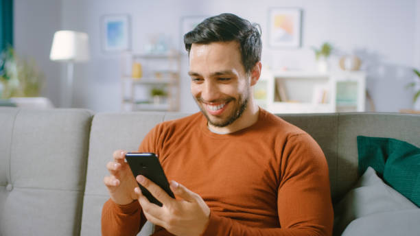 istock Happy Young Man Uses Smartphone while Sitting on a Sofa at Home. Man Browses Through Internet, Watches Videos and Uses Social Networks at Home. 1161176888