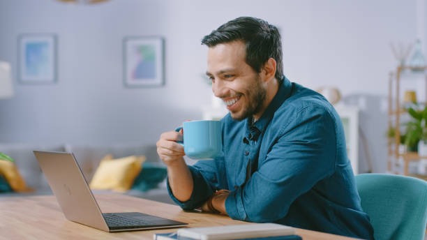 istock Portrait of Handsome Young Man Using Laptop Computer at Home, Watching, Drinking Coffee and Smiling at a Content. Cozy Modern Flat done in Scandinavian Style. 1161176908