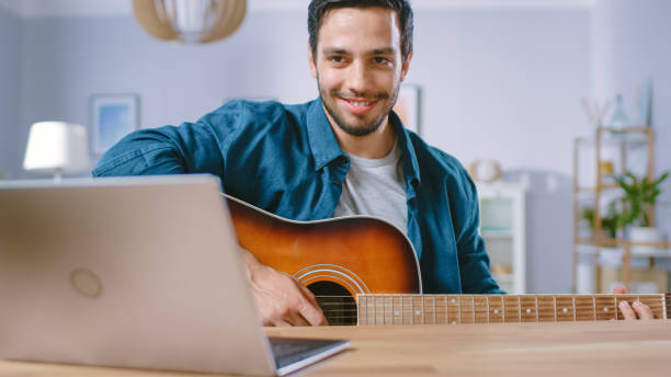 istock Talented Young Man Plays Guitar While Sitting at His Desk, Reading Chords from a Laptop, Doing Online Video Blogging. Man Practicing Guitar Music Playing at Home. 1161176971