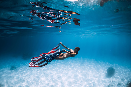 Woman freediver glides over sandy sea bottom with United States flag. Independence day.