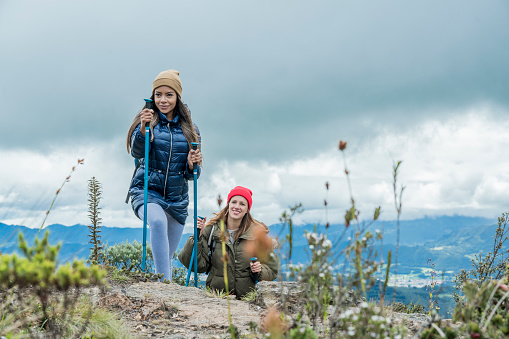 Latin woman on top of the mountain, climbing with the canes in her hand while looking at the camera. Behind her a blond woman walking with the canes in her hand.