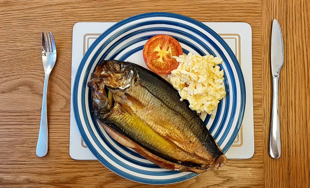 A kipper close up. Whole cold-smoked herring, oily fish, that has been split in a butterfly fashion from tail to head.  Scrambled eggs with half a roasted tomato on a plate with wooden background English breakfast dishes kipper stock pictures, royalty-free photos & images