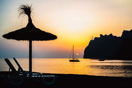 Silhouetted Thatched Sun Shade with Loungers in the Foreground of a Silhouetted Sailboat at Sunrise on the Mediterranean