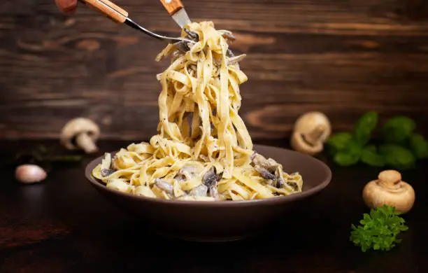 Photo of Homemade Italian fettuccine pasta with mushrooms and cream sauce (Fettuccine al Funghi Porcini). Traditional Italian cuisine. Served on a dark table with a rustic wooden background. Close-up