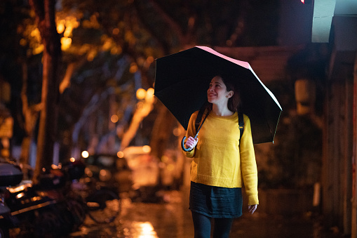 Young , smiling hipster woman walking on the street at night in Shanghai, holding an umbrella