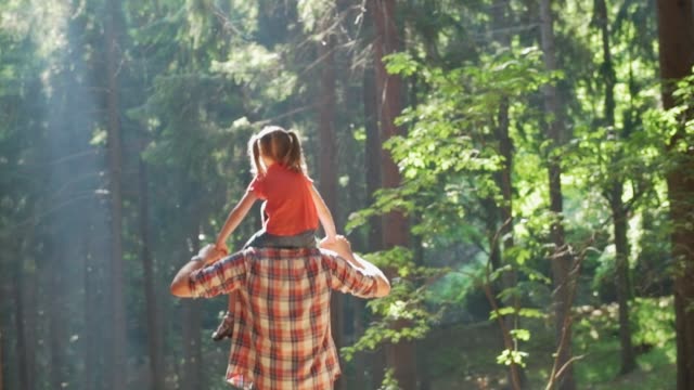 Back view of father giving a piggyback ride to daughter walking in forest.Father holding daughter on shoulders hiking along forest trail path.Father and daughter hiking on forest path together