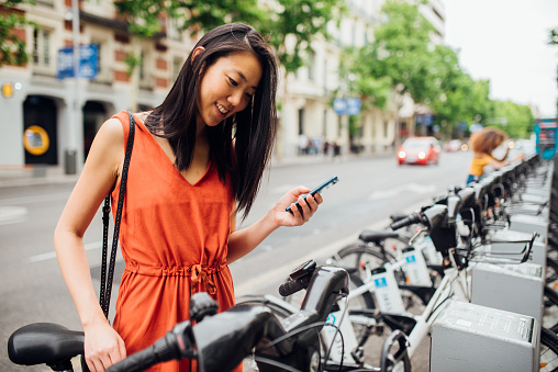 Young cheerful woman taking bicycle out from bicycle rack on the street with smartphone in her hand