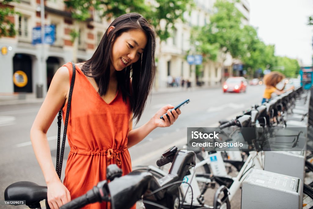 Young woman taking bicycle Young cheerful woman taking bicycle out from bicycle rack on the street with smartphone in her hand Bicycle Sharing System Stock Photo