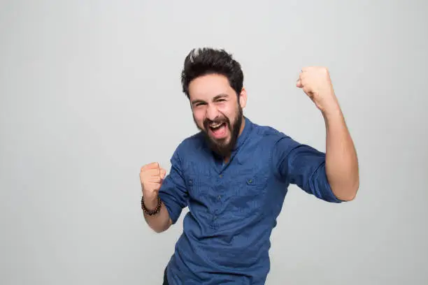 Photo of Portrait of happy young man cheering by shaking fist over grey background