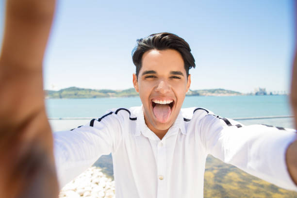Happy Latin man grimacing and taking selfie photo at seaside. Young man showing tongue and holding unseen camera with sea and blue sky in background. Tourism and selfie concept. Front view.