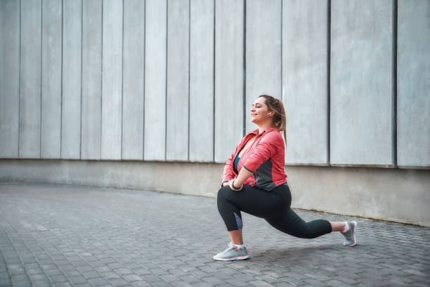 I love sport Happy plus size woman in sport clothes doing stretching exercises and smiling while standing against gray wall. I love sport Happy plus size woman in sport clothes doing stretching exercises and smiling while standing against gray wall. Healthy life. Sport concept. Weight losing. body confidence stock pictures, royalty-free photos & images