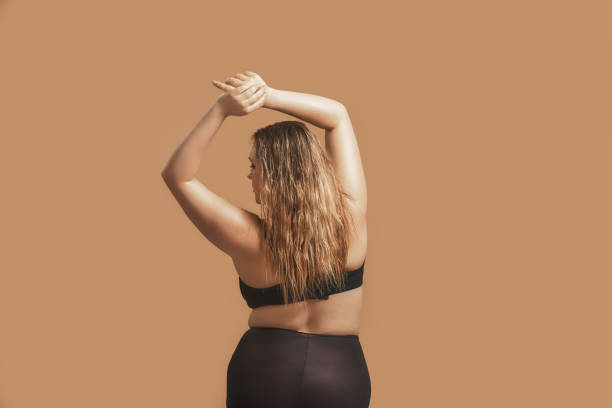 back view of plump sexy woman with long hair in sexy black lingerie raising hands up and posing in studio on brown background - sex symbol sensuality women overweight imagens e fotografias de stock