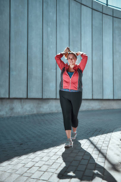 vivre une vie active. pleine longueur de femelle souriante et mignonne de taille plus dans des vêtements de sport ajustant ses cheveux longs et regardant loin tout en restant à l'extérieur - running horses photos et images de collection