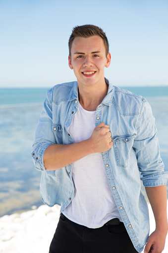 Portrait of positive young man beating chest against seashore. Teenage boy standing at seaside and smiling at camera. Youth culture concept