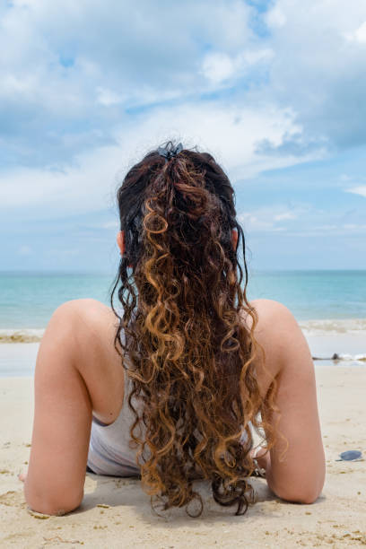 Back view of fair skinned Girl in mid 20s, having curly hairs of golden Color. She is relaxing & sunbathing solo on beach at exotic tropical island. Amazing Back view of fair skinned Girl in mid 20s, having curly hairs of golden Color. She is relaxing & sunbathing solo on beach vacation, to exotic tropical island with clement weather & calm ocean. girls laying on beach stock pictures, royalty-free photos & images