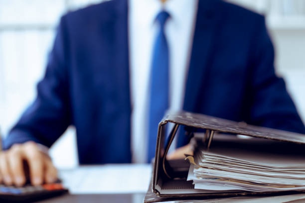 istock Binders with papers waiting to be processed with businessman or bookkeeper back in blur. Accounting planning budget. Audit, insurance and business concept 1161321663