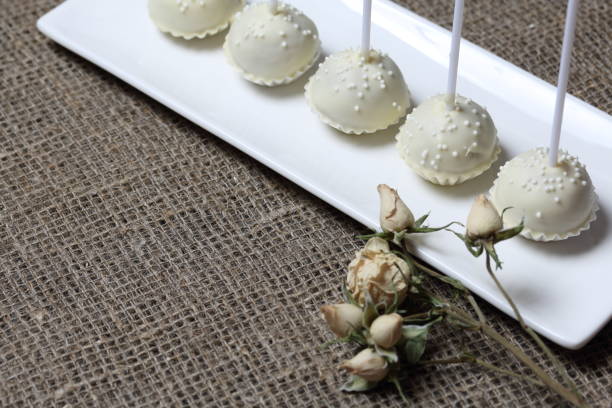 istock Cake pops in white chocolate glaze. Stand vertically on a stand. Decorated with white decorative sprinkles. Near dry beige rose. On a background of coarse linen fabric. 1161327455