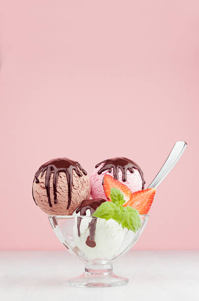 istock Delicious classic different color ice cream scoops in  bowl with mint, spoon, chocolate sauce, strawberry slices in pink interior on white wood board, vertical. 1161336324
