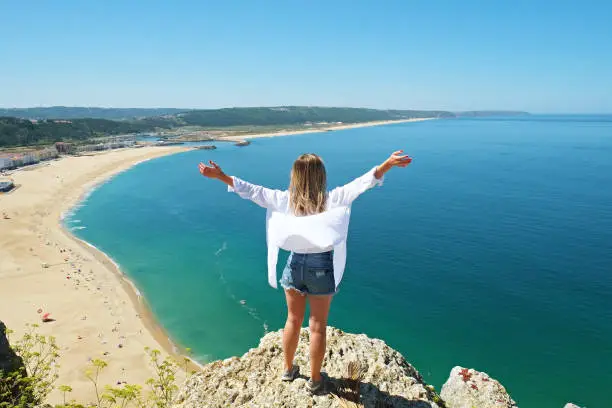 Photo of Young woman wearing oversized cotton shirt enjoying the panoramic top view of famous Nazare beach, Atlantic ocean Algarve, Portugal. Copy space for text, background. Hello summer concept.