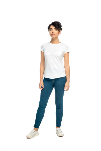 прив�лекательная латинская брюнетка женщина, стоящая в синих джинсах и белой футболке изолированы на белом - полная длина стоковые фото и изображения