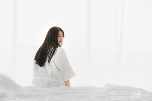Young and beautiful Asian woman sitting on bed in the morning with smiley face and untidy hair after just awake from sleep. Copyspace on the left.