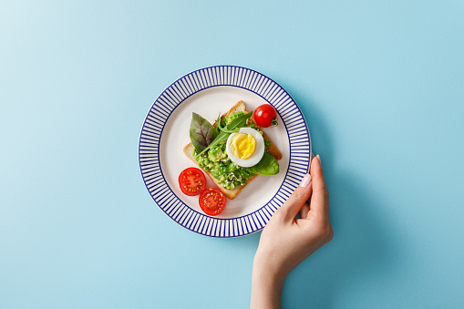 cropped view of woman holding ornamental plate with guacamole, boiled egg and greenery on toast near cherry tomatoes on blue background