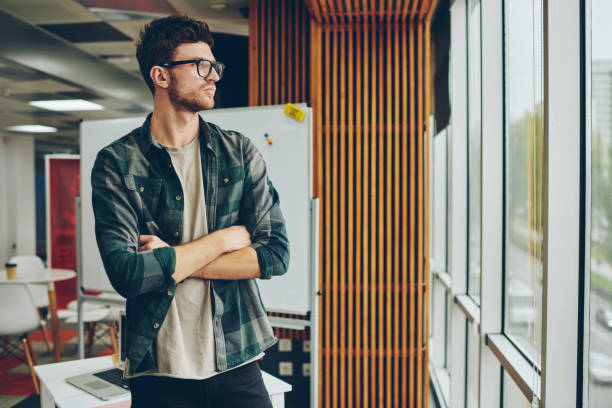 istock Confident young man in eyeglasses for vision correction looking out of window while thinking on development of own startup project.Serious pondering hipster guy with crossed hands 1161356676