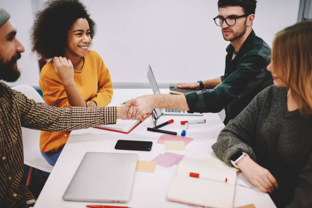 istock Team of creative multicultural designers dressed in formal wear collaborating on developing common project sitting at meeting table.Young men handshaking after successful deal of cooperation 1161357447