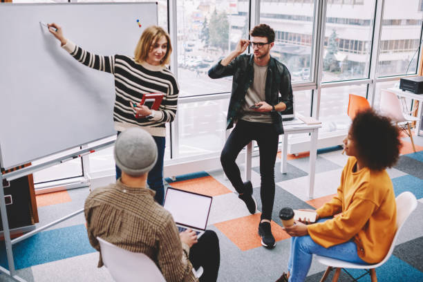 istock Young woman coach pointing on flip chart and explaining studying material group of multicultural students.Professional female teacher conducting workshop team of skilled young people in casual wear 1161357516